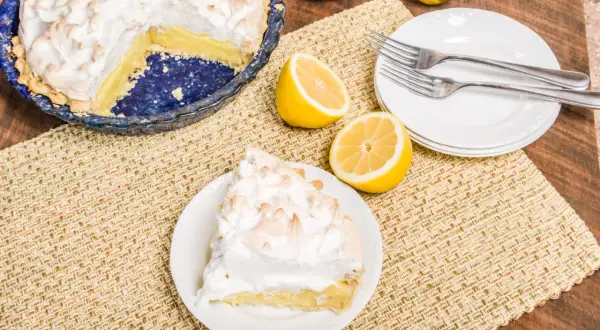 Une part de tarte citron meringuée sur une assiette, à côté d'une tarte entière et de citrons frais. Parfait pour une recette de tarte citron meringuée.
