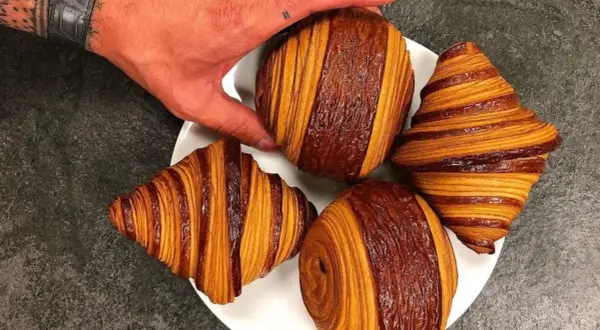 Une main saisit un pain au chocolat Cédric Grolet, aux couches dorées et chocolatées, sur une assiette avec d'autres viennoiseries.