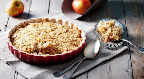 Un délicieux crumble pomme tout juste sorti du four, servi dans un plat rouge, avec une portion sur une cuillère ornée.