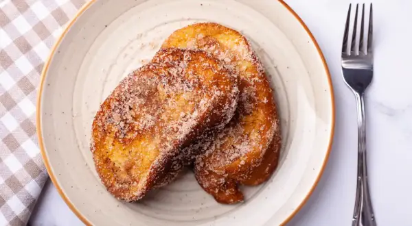 Deux tranches dorées de pain perdu au four, saupoudrées de sucre et de cannelle, sur une assiette avec une serviette à carreaux et une fourchette.