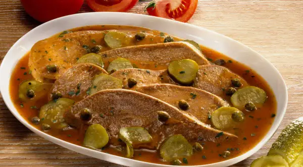 Langue de veau cuisson : fines tranches de langue de veau nappées d'une sauce savoureuse, garnies de cornichons et de câpres. Tomates en arrière-plan.