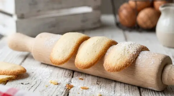 Des biscuits langue de chat dorés, saupoudrés de sucre glace, reposent sur un rouleau à pâtisserie en bois.