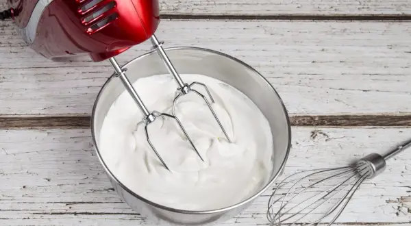 Un batteur rouge prépare une crème chantilly onctueuse dans un bol en inox. Un fouet repose à côté.