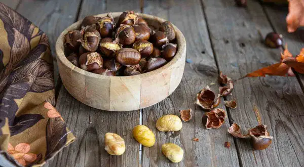 Châtaignes dorées et craquelées, fraîchement sorties de l'air fryer, dans un bol en bois. Quelques châtaignes décortiquées et leurs coquilles éparpillées sur la table en bois.