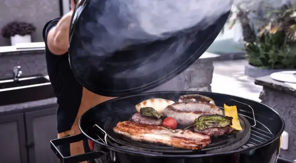 Un homme soulève le couvercle d'un barbecue fumant, révélant des saucisses, des steaks et du bacon. La cuisson lente permet d'attendrir la viande pour un repas savoureux.