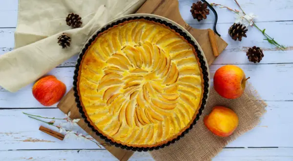 Une tarte aux pommes simple, dorée et appétissante, ornée de fines lamelles de fruits disposées en spirale. Des pommes fraîches, des pommes de pin et une branche de fleurs ajoutent une touche rustique.