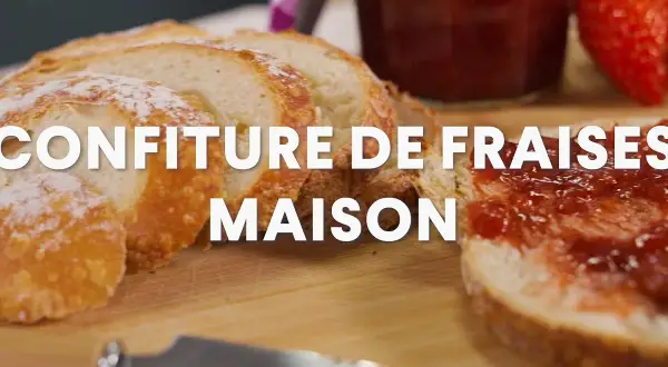 Tranche de pain beurrée et tartinée de confiture de fraise maison, avec une fraise fraîche et un pot de confiture en arrière-plan.