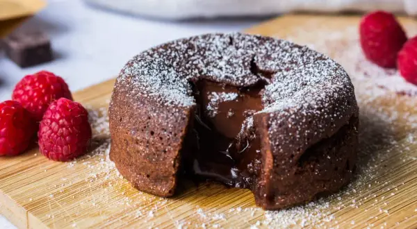 Un fondant au chocolat dégoulinant de cœur coulant, saupoudré de sucre glace, avec des framboises fraîches sur une planche en bois.