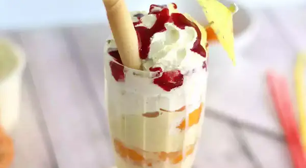Un déliceux pêche Melba, avec des couches de fruits, de crème fouettée et de coulis de fruits rouges, garni d'une gaufrette et d'un petit parasol.