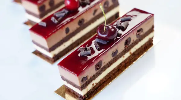 Délicieux entremets Forêt Noire, une symphonie chocolatée et cerise, parsemée de copeaux de chocolat et surmontée d'une cerise fraîche.
