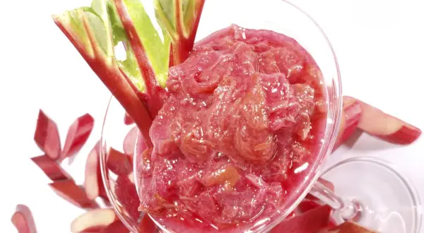 Un verre rempli de compote de rhubarbe, garni de tiges fraîches. Des morceaux de rhubarbe coupés entourent le verre.