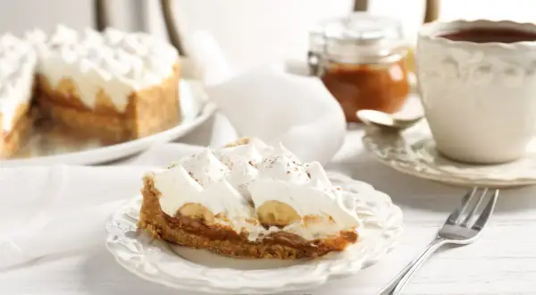 Tranche de tarte bananes et crème fouettée, avec une touche de confiture de lait. Une tasse de thé et une petite jarre complètent ce tableau gourmand.