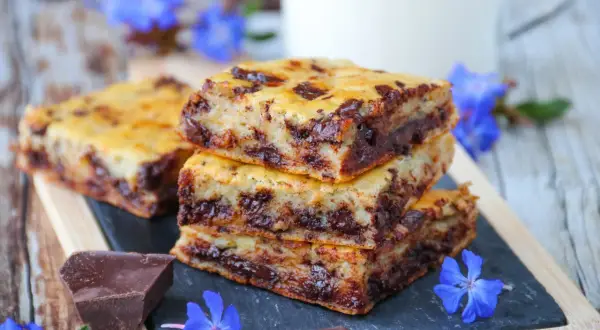 Gourmandises de cake banane pépite chocolat, empilées sur une ardoise, décorées de fleurs bleues et de morceaux de chocolat noir.
