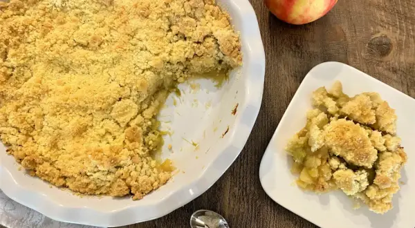 Un délicieux crumble pomme rhubarbe, doré et croustillant, servi dans un plat blanc avec une portion sur une assiette. Une pomme rouge et jaune repose à côté.