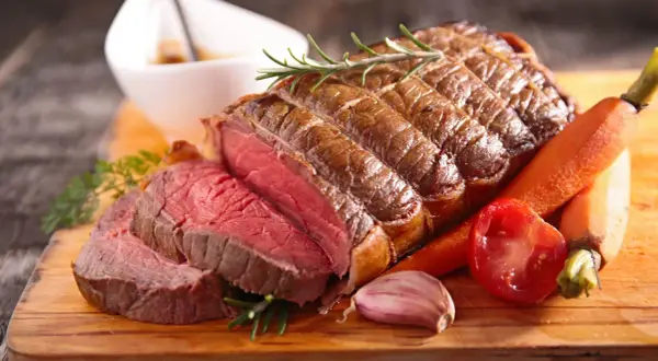 Un rôti de bœuf congelé au four, tranché, avec des carottes, une tomate, de l'ail et du romarin sur une planche en bois.