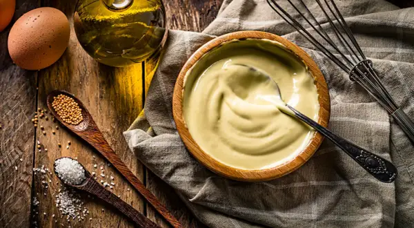 Un bol de mayonnaise maison crémeuse, avec des ingrédients frais : œuf, huile, moutarde et sel. Un fouet à portée de main.