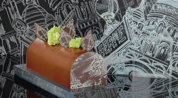 Bûche de Noël chocolatée, décorée de chocolat blanc et de mousse verte, évoquant une recette Pierre Hermé. Le fond présente des croquis architecturaux parisiens.