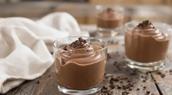Trois verrines de mousse au chocolat Alain Ducasse, garnies de copeaux de chocolat, sur une table en bois rustique.