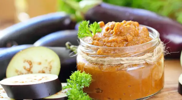 Un pot de caviar d'aubergine, garni de persil frais, entouré d'aubergines entières et tranchées.