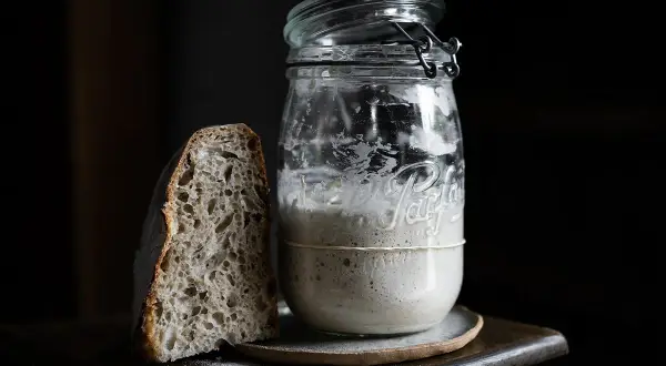 Une tranche de pain au levain maison à côté d'un bocal rempli de levain actif.