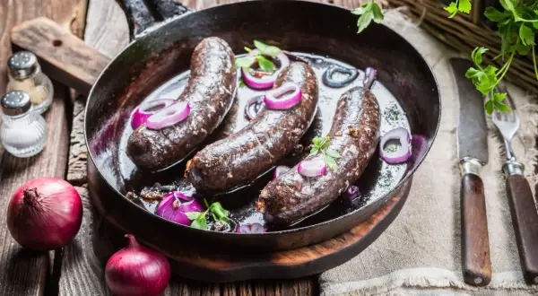 Trois boudins noirs en cuisson dans une poêle, accompagnés d'oignons rouges et de persil. Un plat rustique prêt à être dégusté.
