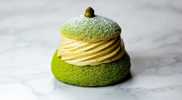 Un Paris Brest pistache, un délice vert et doré, saupoudré de sucre glace, sur fond marbré.