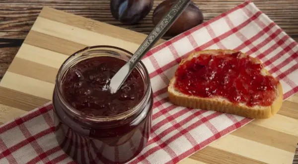 Un pot de confitures de quetsches maison, une tartine généreusement garnie et quelques quetsches sur une planche en bois.