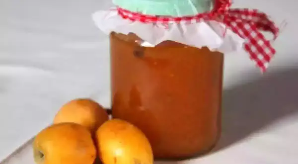 Un pot de confiture de nèfles, décoré d'un ruban à carreaux rouges et blancs, est posé à côté de trois fruits frais.