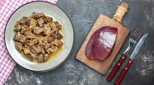 Un plat de foie de veau cuisiné avec des oignons, prêt à être dégusté. À côté, une tranche de foie de veau crue sur une planche, avec des couverts.