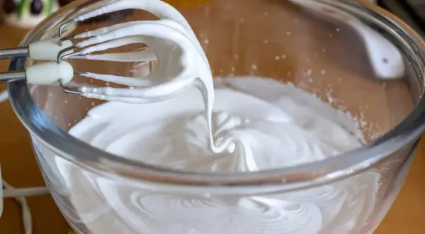 Fouets de batteur électrique en train de battre les blancs en neige dans un bol en verre, créant une texture aérienne parfaite pour une meringue.