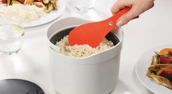 Une spatule orange sert du riz rond dans un cuiseur blanc. Le riz est prêt pour un repas savoureux.