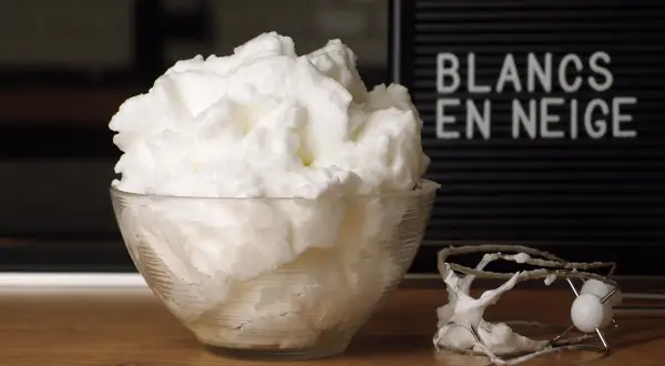 Un bol de blancs en neige aériens, comme un nuage, à côté d'un batteur. Le mot "BLANCS EN NEIGE" est visible sur un panneau.