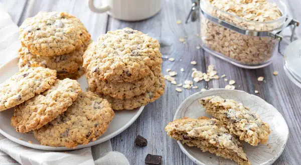 Des biscuits flocons d'avoine aux pépites de chocolat, empilés sur une assiette, avec un bocal de flocons d'avoine en arrière-plan.