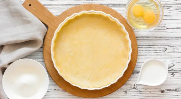 Une pâte à tarte crue est prête dans un plat blanc, entourée d'ingrédients pour différentes pâtes à tarte : œufs, lait, farine.