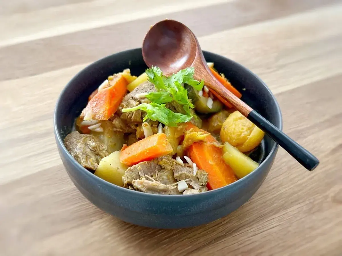 Un délicieux tajine agneau aux carottes et pommes de terre, garni de coriandre fraîche. Une cuillère en bois repose sur le plat.