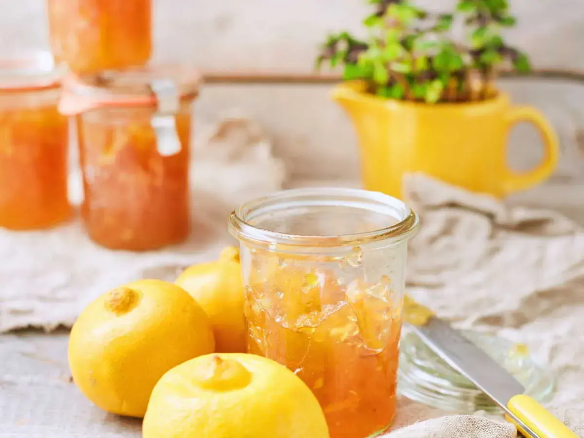 Bocaux de marmelade de citron italienne, avec des citrons frais et une plante verte dans un pichet jaune en arrière-plan.