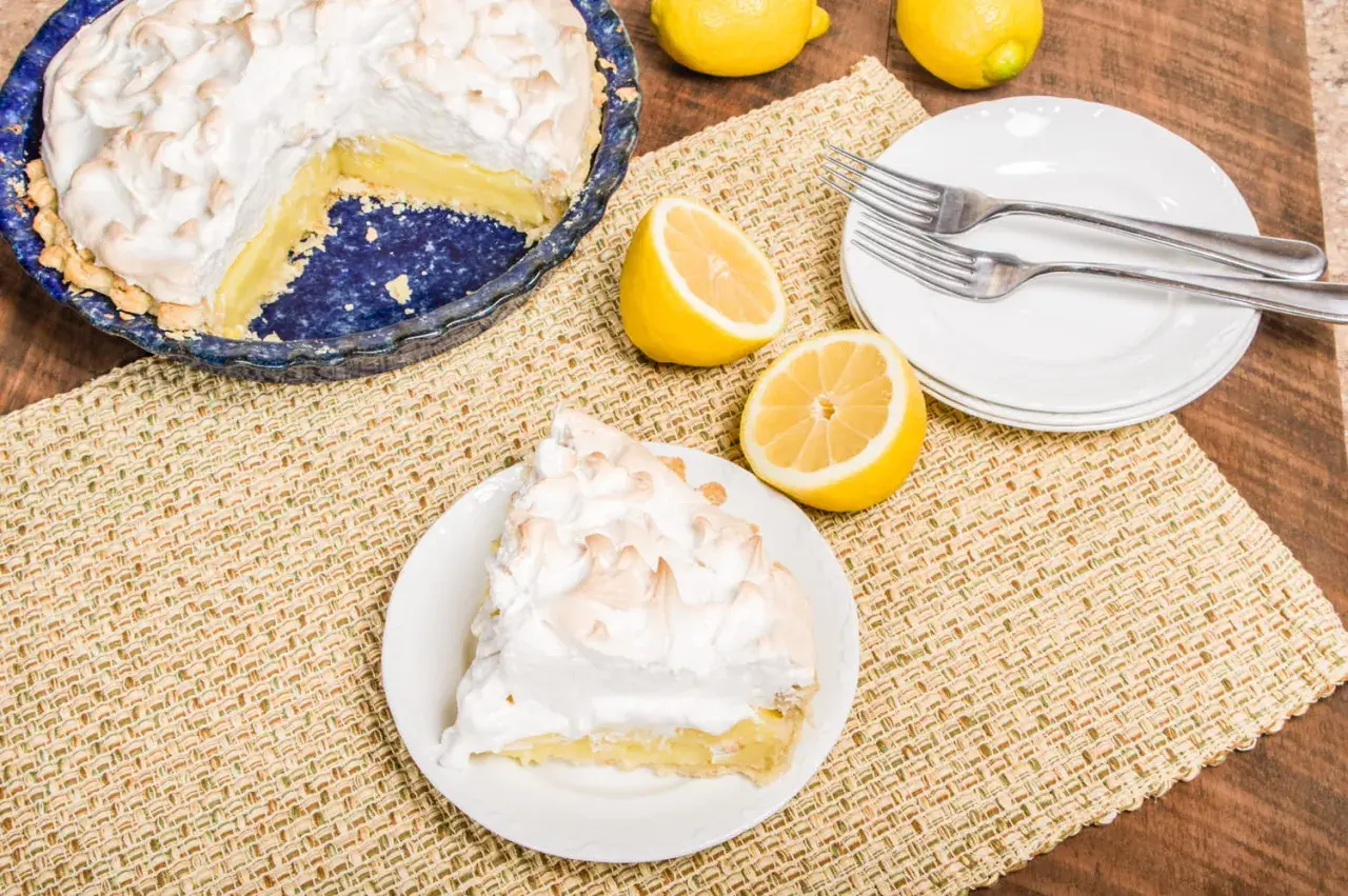 Une part de tarte citron meringuée sur une assiette, à côté d'une tarte entière et de citrons frais. Parfait pour une recette de tarte citron meringuée.