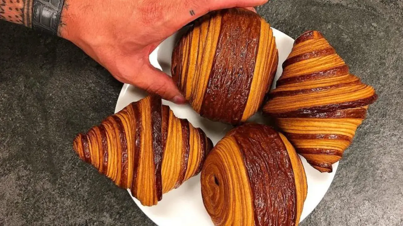 Une main saisit un pain au chocolat Cédric Grolet, aux couches dorées et chocolatées, sur une assiette avec d'autres viennoiseries.