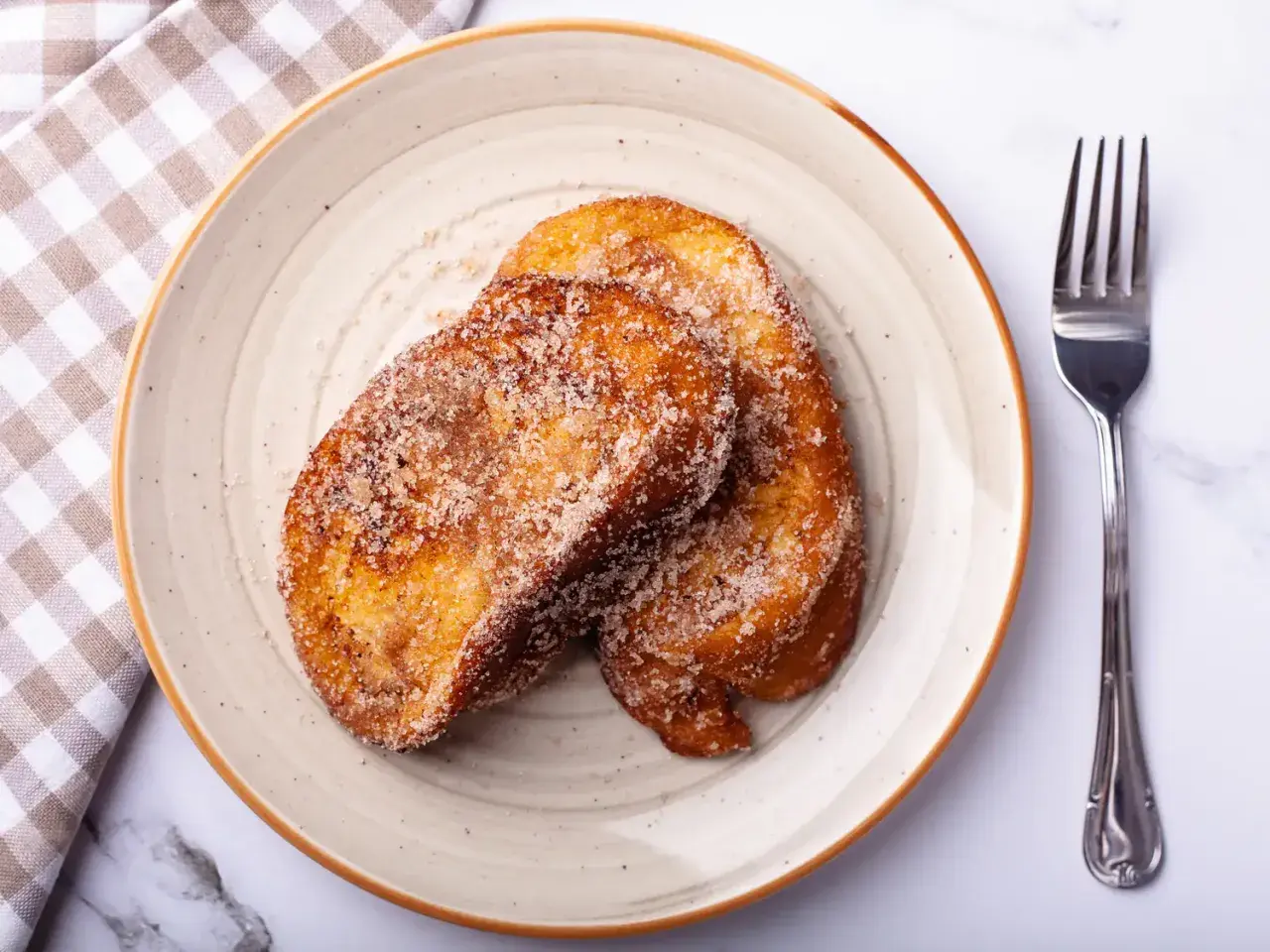 Deux tranches dorées de pain perdu au four, saupoudrées de sucre et de cannelle, sur une assiette avec une serviette à carreaux et une fourchette.