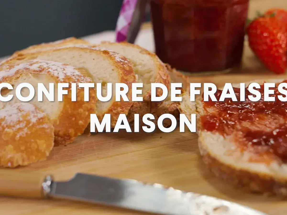 Tranche de pain beurrée et tartinée de confiture de fraise maison, avec une fraise fraîche et un pot de confiture en arrière-plan.