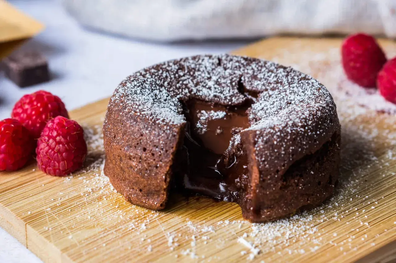 Un fondant au chocolat dégoulinant de cœur coulant, saupoudré de sucre glace, avec des framboises fraîches sur une planche en bois.