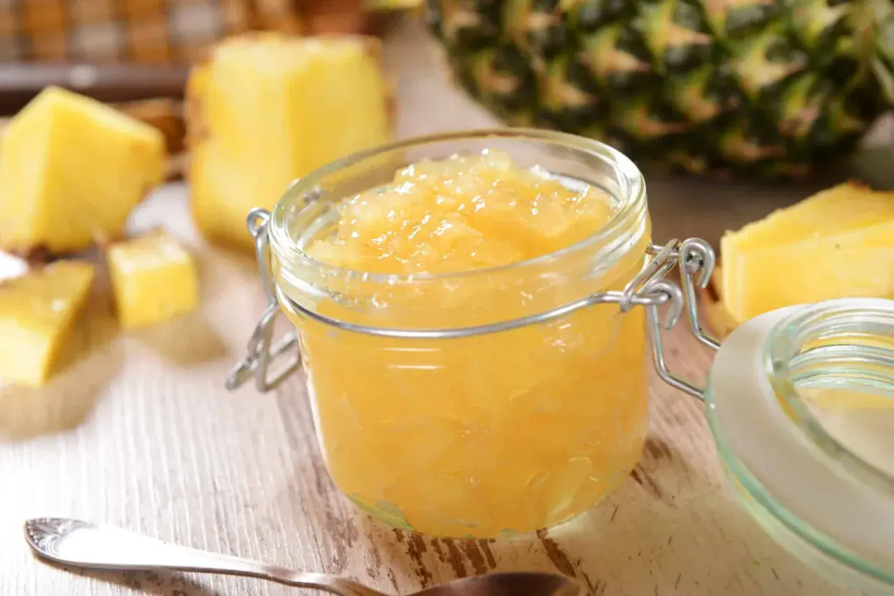 Un pot de confiture d'ananas maison, entouré de morceaux d'ananas frais et d'une cuillère.