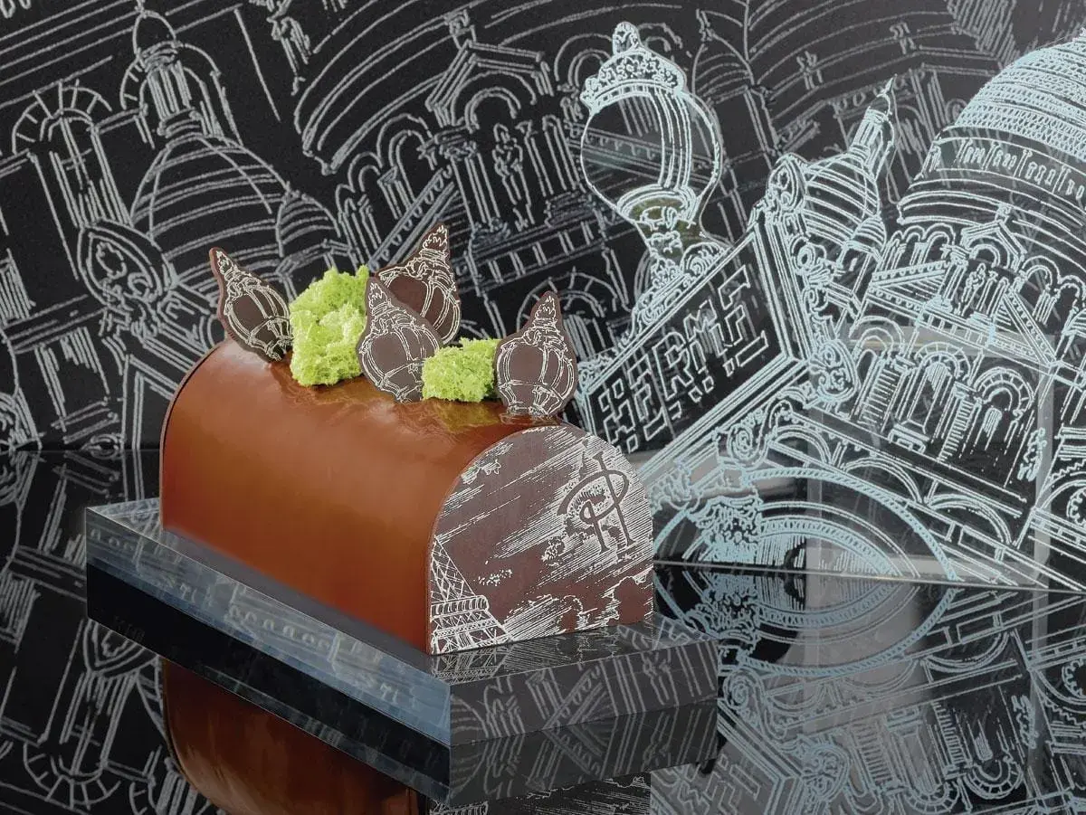 Bûche de Noël chocolatée, décorée de chocolat blanc et de mousse verte, évoquant une recette Pierre Hermé. Le fond présente des croquis architecturaux parisiens.