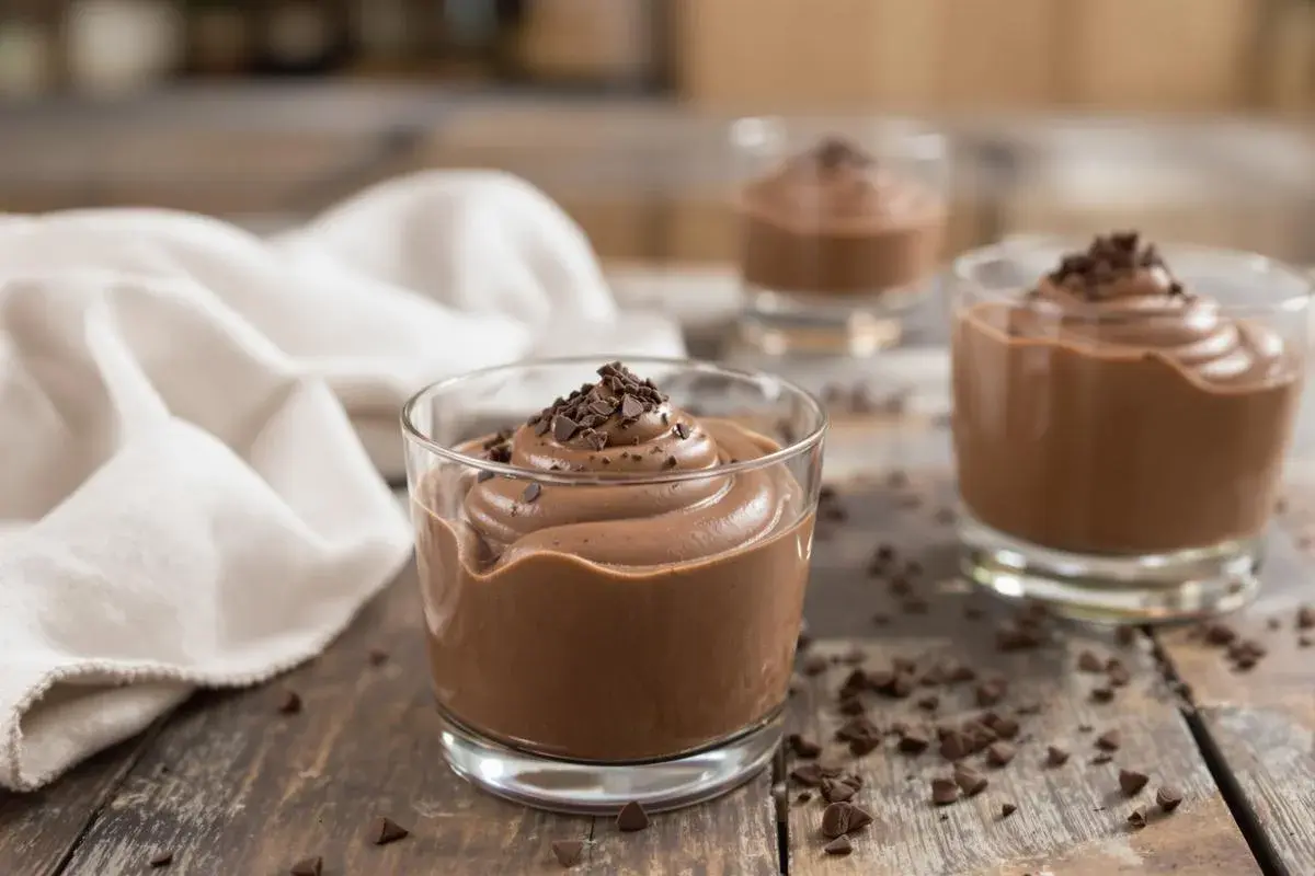 Trois verrines de mousse au chocolat Alain Ducasse, garnies de copeaux de chocolat, sur une table en bois rustique.