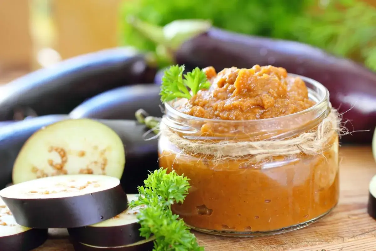 Un pot de caviar d'aubergine, garni de persil frais, entouré d'aubergines entières et tranchées.
