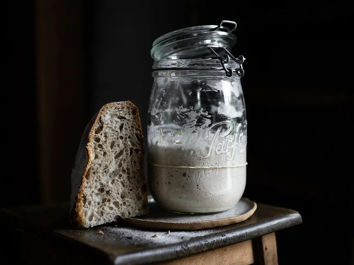 Une tranche de pain au levain maison à côté d'un bocal rempli de levain actif.