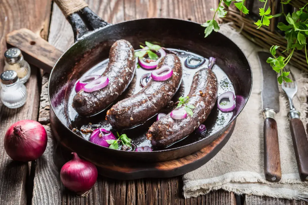 Trois boudins noirs en cuisson dans une poêle, accompagnés d'oignons rouges et de persil. Un plat rustique prêt à être dégusté.