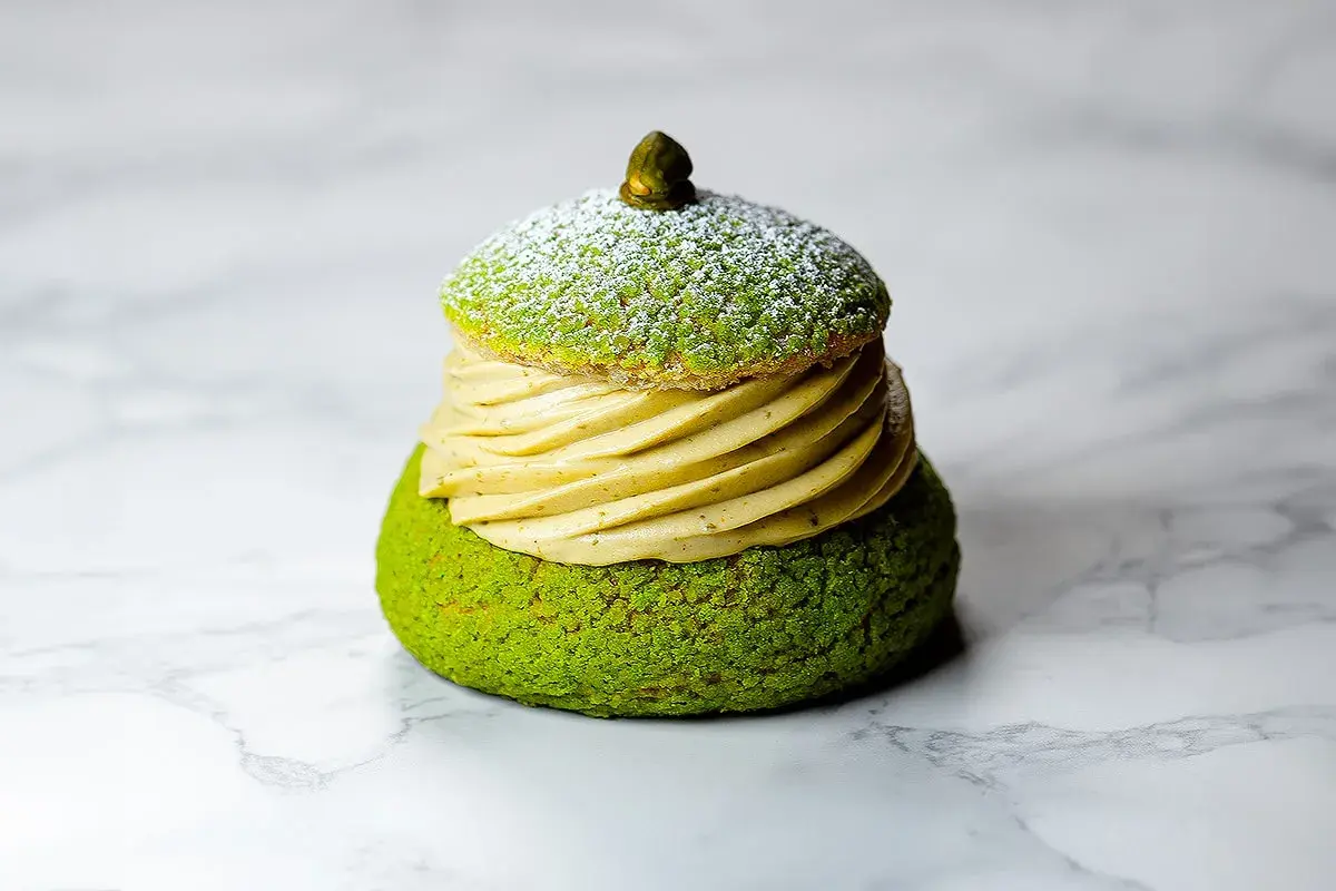 Un Paris Brest pistache, un délice vert et doré, saupoudré de sucre glace, sur fond marbré.