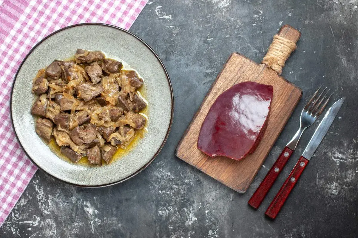 Un plat de foie de veau cuisiné avec des oignons, prêt à être dégusté. À côté, une tranche de foie de veau crue sur une planche, avec des couverts.