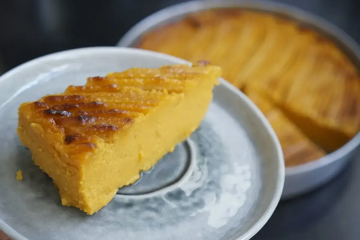 Tranche de gâteau patate douce, dorée et moelleuse, servie sur une assiette grise. Le reste du gâteau est visible en arrière-plan.